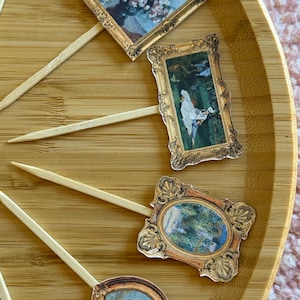 May include: A collection of miniature framed art prints attached to wooden skewers, arranged on a bamboo tray. The art features classic paintings with gold-colored frames. The paintings depict floral arrangements and landscapes.