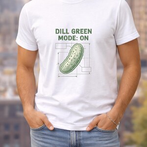 Peut inclure: T-shirt blanc avec l'inscription "DILL GREEN MODE: ON" au-dessus d'une illustration de cornichon vert. Le cornichon est détaillé avec un design de type plan. Le t-shirt est porté par une personne en jean bleu.