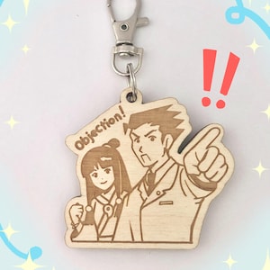 May include: Wooden keychain featuring a cartoon illustration of two characters with the text "Objection!". The keychain has a silver clasp and a light wood finish. The characters are a man pointing and a woman.