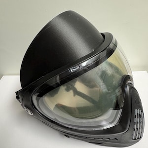 Peut inclure: Masque de paintball noir avec un objectif clair et incurvé. Le masque a une visière noire et une conception protectrice autour du visage. L'objectif reflète l'environnement. Le masque est conçu pour la protection lors des jeux de paintball.