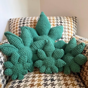May include: A collection of handmade, green, leaf-shaped throw pillows. The pillows are arranged on a patterned armchair. The leaves have a textured, knitted appearance, and vary in size. The color is a vibrant, medium green.
