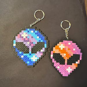 Könnte beinhalten: Zwei außerirdisch geformte Schlüsselanhänger aus bunten Perlen. Einer hat ein blaues, lila und weißes Galaxiemuster, der andere zeigt rosa, orange und lila Farbtöne. Beide haben schwarze Augen- und Munddetails und silberne Schlüsselringe.
