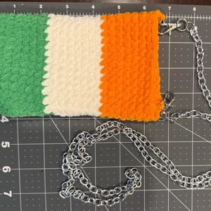 Può includere: Una borsa a tracolla all'uncinetto nei colori della bandiera irlandese: verde, bianco e arancione. La borsa ha una tracolla a catena argentata e chiusure argentate. La borsa è rettangolare e ha un aspetto strutturato.