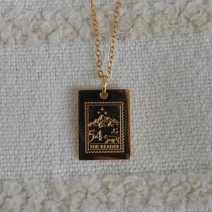 May include: A gold-colored necklace with a rectangular pendant. The pendant features a mountain scene with the text "54" and "THE LEADER" in a vintage stamp design. The necklace hangs on a white textured surface.