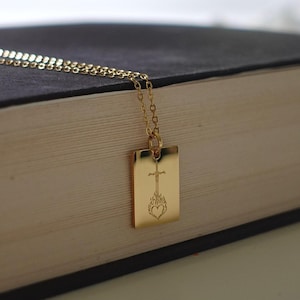 Puede incluir: Un collar dorado con un colgante rectangular con un diseño de cruz sobre un corazón. El collar está sobre un libro oscuro, con un fondo borroso.