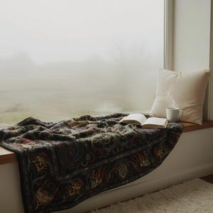 May include: A cozy window seat scene featuring a patterned blanket, an open book, a white pillow, and a mug. The blanket has a dark background with ornate, colorful designs. The scene is bathed in soft, natural light.