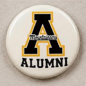 May include: A circular pinback button featuring a white background with a large black letter A outlined in yellow. The word Mountaineers is written in black script across the center of the letter A, and the word ALUMNI is printed in black below.