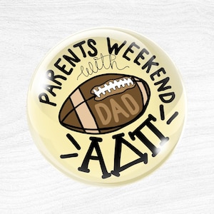 Pode incluir: Um botão redondo com fundo creme apresentando uma ilustração de uma bola de futebol americano marrom no centro. O texto diz PARENTS WEEKEND with DAD em letras marrons, e as letras gregas Alpha Delta Pi estão impressas em preto abaixo.