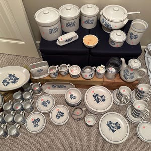 May include: A collection of white ceramic kitchenware and tableware with blue floral designs. Includes canisters, serving dishes, plates, cups, saucers, a teapot, and a butter dish. The set is arranged on a dark blue ottoman and a wooden surface.