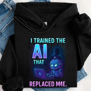 Może przedstawiać: Czarna bluza z kapturem z napisem "I TRAINED THE AI THAT REPLACED ME." Projekt przedstawia kreskówkowego robota pracującego na laptopie, na tle obwodu drukowanego. Tekst i robot są w odcieniach niebieskiego i fioletowego.