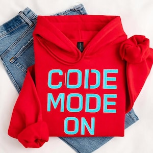 Pode incluir: Um moletom vermelho com a frase "CODE MODE ON" em letras azul claro. O moletom está dobrado e colocado sobre um par de jeans azuis. A peça tem capuz e mangas compridas.