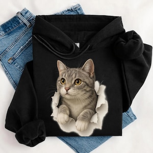 May include: Black hoodie featuring a realistic graphic of a gray tabby cat emerging from a torn white paper effect. The cat has yellow eyes and is looking forward. The hoodie has a drawstring hood and long sleeves.