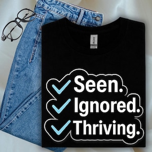 Könnte beinhalten: Schwarzes T-Shirt mit dem Text "Seen. Ignored. Thriving." in Weiß, umrandet von einer Wolkenform, mit blauen Häkchen. Das Shirt wird mit einer blauen Jeans und einer schwarzen Brille kombiniert.