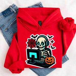 May include: A red hoodie featuring a Halloween design. The graphic shows a skeleton wearing headphones and using a computer, alongside a jack-o'-lantern, bats, and a spiderweb. The hoodie is displayed with blue jeans.