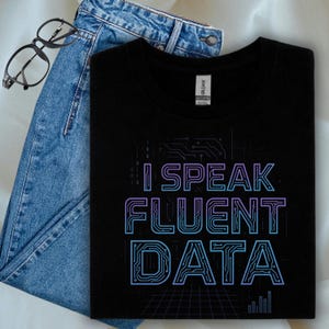 Puede incluir: Camiseta negra con el texto "I Speak Fluent Data" en un diseño de placa de circuito, con tonos azules y morados. La camiseta se combina con vaqueros azules y gafas con montura negra.
