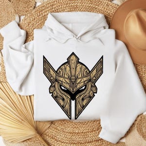Puede incluir: Sudadera con capucha blanca con una ilustración detallada de un casco de guerrero en tonos dorados y negros. El diseño está centrado en la parte delantera. El casco presenta patrones intrincados y una expresión severa. La sudadera tiene una capucha con cordón.