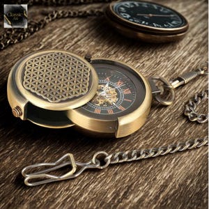Op de afbeelding: Antieke messing zakhorloge met een gedetailleerde open wijzerplaat, Romeinse cijfers en een decoratief geometrisch patroon op de deksel. Het horloge wordt vergezeld door een ketting en een sluiting, op een houten oppervlak.