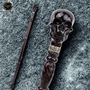 Pode incluir: Bengala castanha escura com uma pega em forma de caveira. A pega tem uma faixa prateada com um padrão decorativo. O eixo é fino com detalhes dourados. O design geral da bengala é gótico.