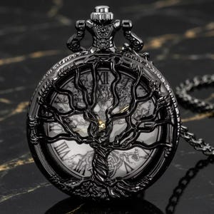 Puede incluir: Un reloj de bolsillo negro con un diseño recortado de árbol en la tapa. La esfera muestra números romanos sobre un fondo blanco con una ilustración de un árbol. Incluye una cadena negra a juego sujeta a la corona superior.