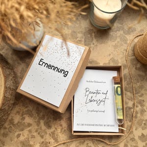 May include: A gift box with the word "Ernennung" printed on the lid. Inside, a card reads "Beamten auf Lebenszeit" and a small glass tube containing a 10 Euro bill. The box is brown with a white interior.