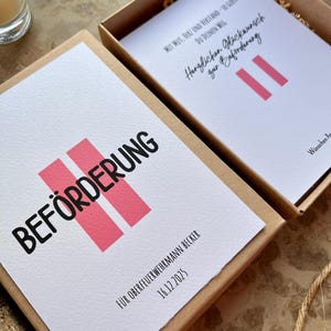 May include: A gift box with a white card featuring the word "BEF&Ouml;RDERUNG" in black, with two pink vertical bars. The text "F&Uuml;R OBERFEUERWEHRMANN BECKER 16.12.2025" is also visible. The box is open, revealing another card.