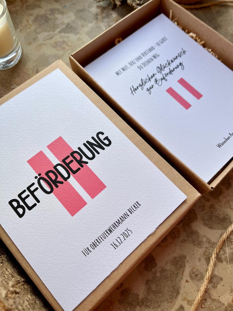 May include: A gift box with a white card featuring the word "BEF&Ouml;RDERUNG" in black, with two pink vertical bars. The text "F&Uuml;R OBERFEUERWEHRMANN BECKER 16.12.2025" is also visible. The box is open, revealing another card.