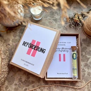 May include: A gift box with a white card that reads "BEF&Ouml;RDERUNG" in black and pink. Inside the box is a card with text and a glass vial containing a Euro bill. The box is on a textured surface with decorative elements.