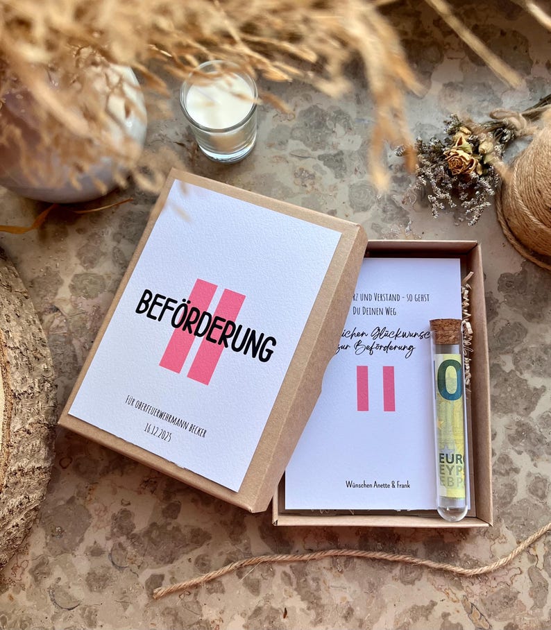 May include: A gift box with a white card that reads "BEF&Ouml;RDERUNG" in black and pink. Inside the box is a card with text and a glass vial containing a Euro bill. The box is on a textured surface with decorative elements.