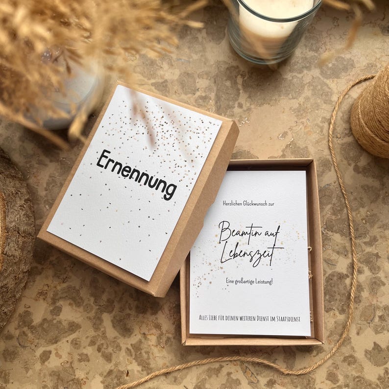 May include: A small, open cardboard box with a white card inside. The card reads "Beamten auf Lebenszeit" in elegant script. The box lid, also with a white card, displays the word "Ernennung". Gold flecks and a candle are visible.