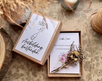 May include: A small, open cardboard gift box with a white card inside and a decorative dried flower bouquet. The top card reads "YAAAY! Ausbildung bestanden" with a champagne bottle illustration.