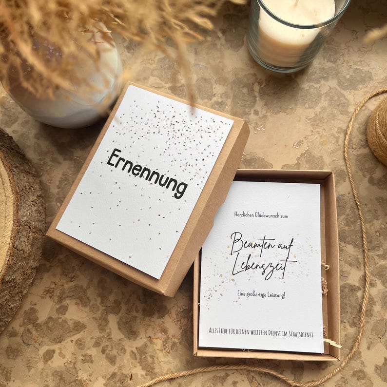 May include: A brown cardboard gift box with a white card inside. The card reads "Ernennung" in black text. The box is open, revealing another card with script. The image includes a candle in a glass and decorative elements.