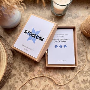May include: A gift box with a white card inside and a lid with the word "BEF&Ouml;RDERUNG" in black and a blue star design. The box is open, revealing a card with text and blue stars. A candle and twine are in the background.