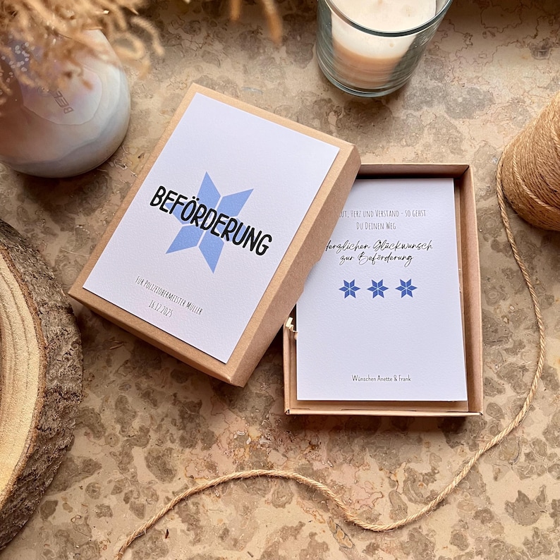 May include: A gift box with a white card inside and a lid with the word "BEF&Ouml;RDERUNG" in black and a blue star design. The box is open, revealing a card with text and blue stars. A candle and twine are in the background.