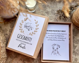May include: A graduation gift box with a card that says "GESCHAFFT!" and "Paula". The box is open, revealing a card with text in German. A small candle and decorative elements are in the background.
