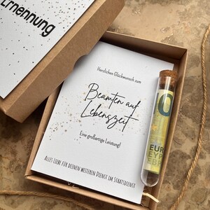 May include: A gift box with a greeting card and a glass vial. The card reads "Herzlichen Gl&uuml;ckwunsch zum Beamten auf Lebenszeit" in elegant script. A ten Euro bill is inside the vial. The box is brown.