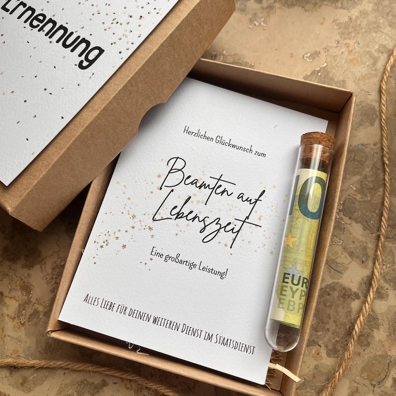 May include: A gift box with a greeting card and a glass vial. The card reads "Herzlichen Gl&uuml;ckwunsch zum Beamten auf Lebenszeit" in elegant script. A ten Euro bill is inside the vial. The box is brown.
