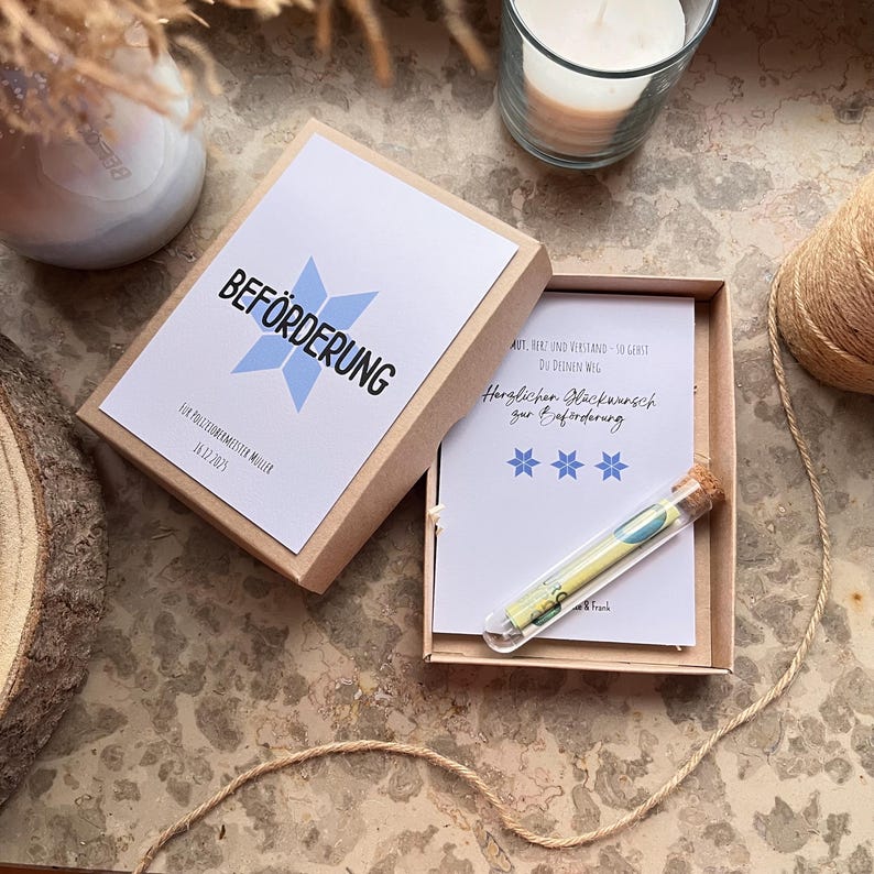 May include: A gift box with the word "BEF&Ouml;RDERUNG" in black text and a light blue starburst design. Inside, a card with text and a small glass tube with a rolled-up item. The box is open, revealing its contents.