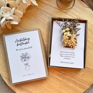 Könnte beinhalten: Zwei offene Geschenkboxen mit Karten und Trockenblumen. Eine Karte lautet "Ausbildung bestanden" und "Wir sind so stolz auf dich!" mit einer Blumenstrauß-Illustration und "Für Amelie". Die andere Karte hat Text auf Deutsch und ein Trockenblumenarrangement.