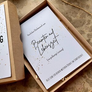 May include: A gift box with a white card inside. The card has elegant script that reads "Beamten auf Lebenszeit" with the text "Herzlichen Gl&uuml;ckwunsch zum" above and "Eine gro&szlig;artige Leistung!" below. The box is brown.