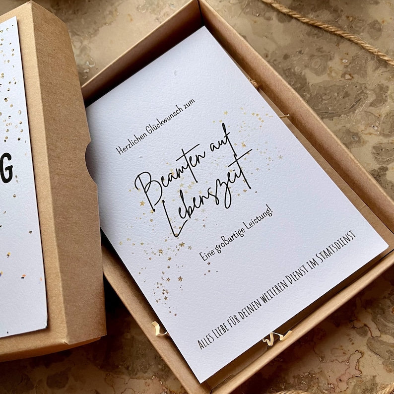 May include: A gift box with a white card inside. The card has elegant script that reads "Beamten auf Lebenszeit" with the text "Herzlichen Gl&uuml;ckwunsch zum" above and "Eine gro&szlig;artige Leistung!" below. The box is brown.