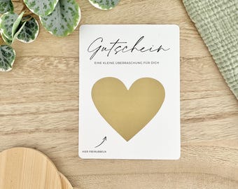 May include: A white gift certificate with the word "Gutschein" in elegant script. It features a gold heart-shaped scratch-off area and the text "EINE KLEINE ÜBERRASCHUNG FÜR DICH" and "HIER FREIRUBBELN".