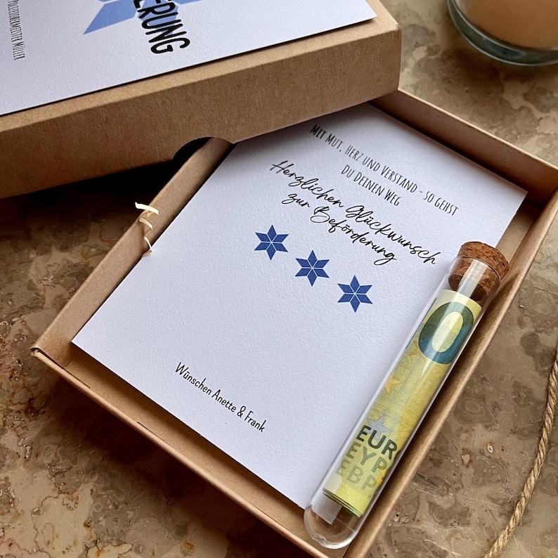 May include: A gift box with a greeting card and a glass vial. The card has text and blue star designs. The vial contains a rolled-up Euro bill. The box is brown, and the card is white.