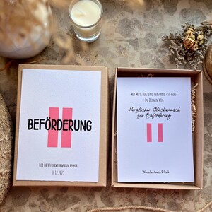 May include: A gift box with a white card that says "BEF&Ouml;RDERUNG" in black letters with two pink vertical bars. Below the text is "F&Uuml;R OBERFEUERWEHRMANN BECKER 16.12.2025". The box is open, revealing another card with German text and pink bars.