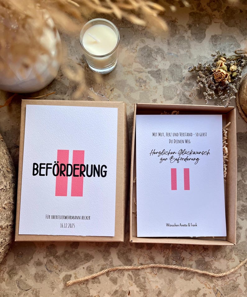 May include: A gift box with a white card that says "BEF&Ouml;RDERUNG" in black letters with two pink vertical bars. Below the text is "F&Uuml;R OBERFEUERWEHRMANN BECKER 16.12.2025". The box is open, revealing another card with German text and pink bars.