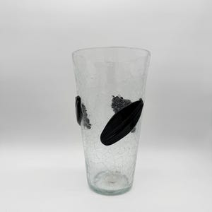 May include: A clear glass vase featuring a distinct crackle texture throughout the body. The vase has a tapered, cylindrical shape and is decorated with two smooth, solid black, leaf-shaped glass appliques attached to the exterior surface.