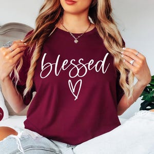 May include: Maroon t-shirt with the word "blessed" in white script and a white heart graphic. The shirt is a solid color and has a round neckline. The model is wearing ripped blue jeans.