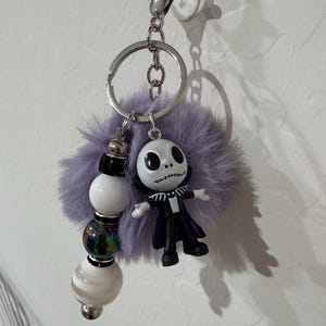 May include: A silver keychain with a Jack Skellington figure, a fluffy purple pom-pom, and decorative beads. The Jack Skellington figure is wearing a black suit and has a stitched smile. The beads are white, iridescent, and striped.