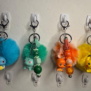 May include: Four keychains with cartoon character charms, fluffy pom-poms, and beaded accents. The keychains feature a blue Squirtle, a green Bulbasaur, an orange Charmander, and a yellow Pikachu. Each keychain has a silver clasp and a tassel.