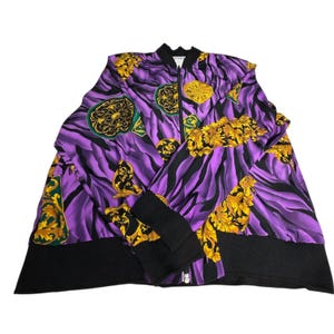 May include: A purple and black patterned bomber jacket with gold accents. The jacket features a black ribbed collar, cuffs, and hem, and a full-length zipper. The design includes a zebra stripe pattern with gold baroque-style embellishments.