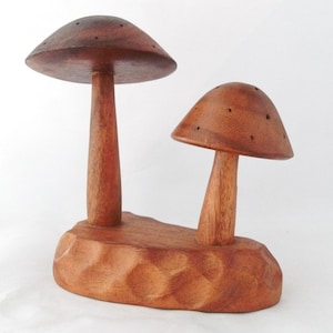May include: A wooden mushroom-shaped sculpture with two mushroom caps of different sizes. The sculpture is carved from a reddish-brown wood and sits on a textured base. The caps have small holes.
